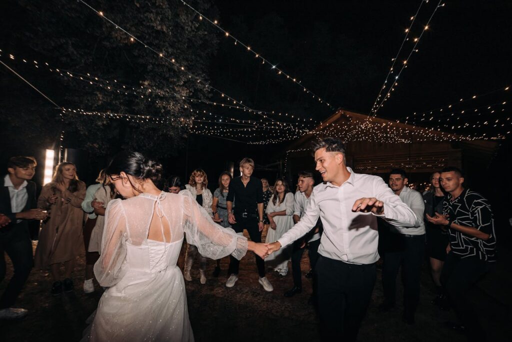 A couple dances joyfully under twinkling lights at an outdoor night wedding celebration.